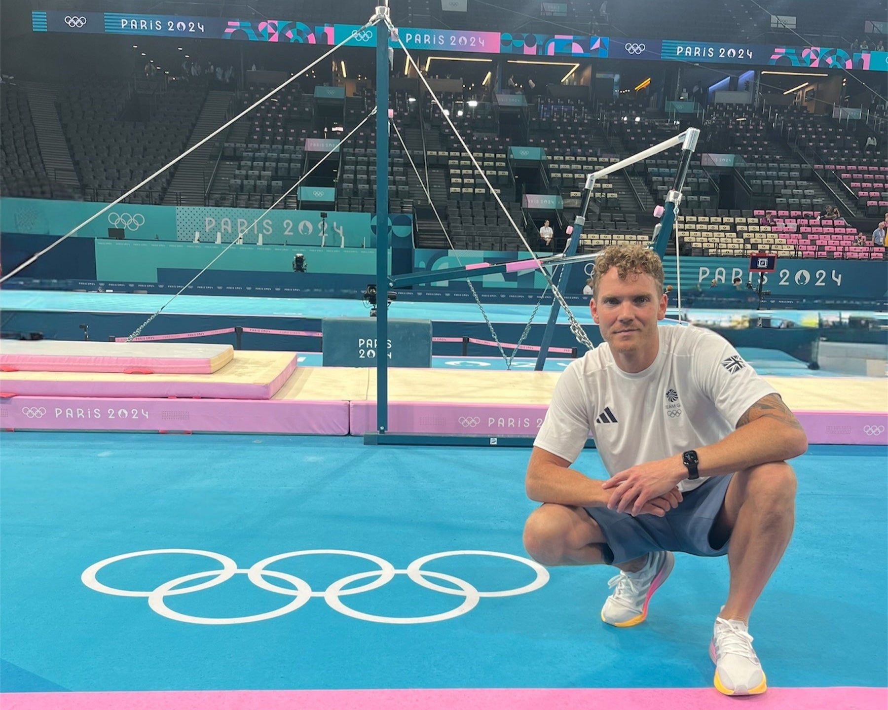 Man posing on a gymnastics floor with Olympic rings logo in the background